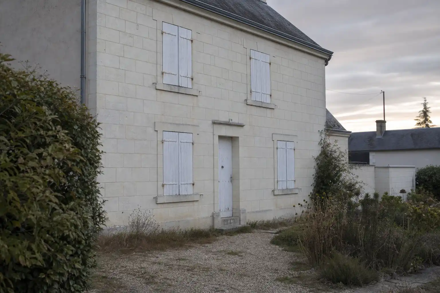 Maison de bourg en tuffeau aux volets de bois fermés, allée de gravier et jardin sec